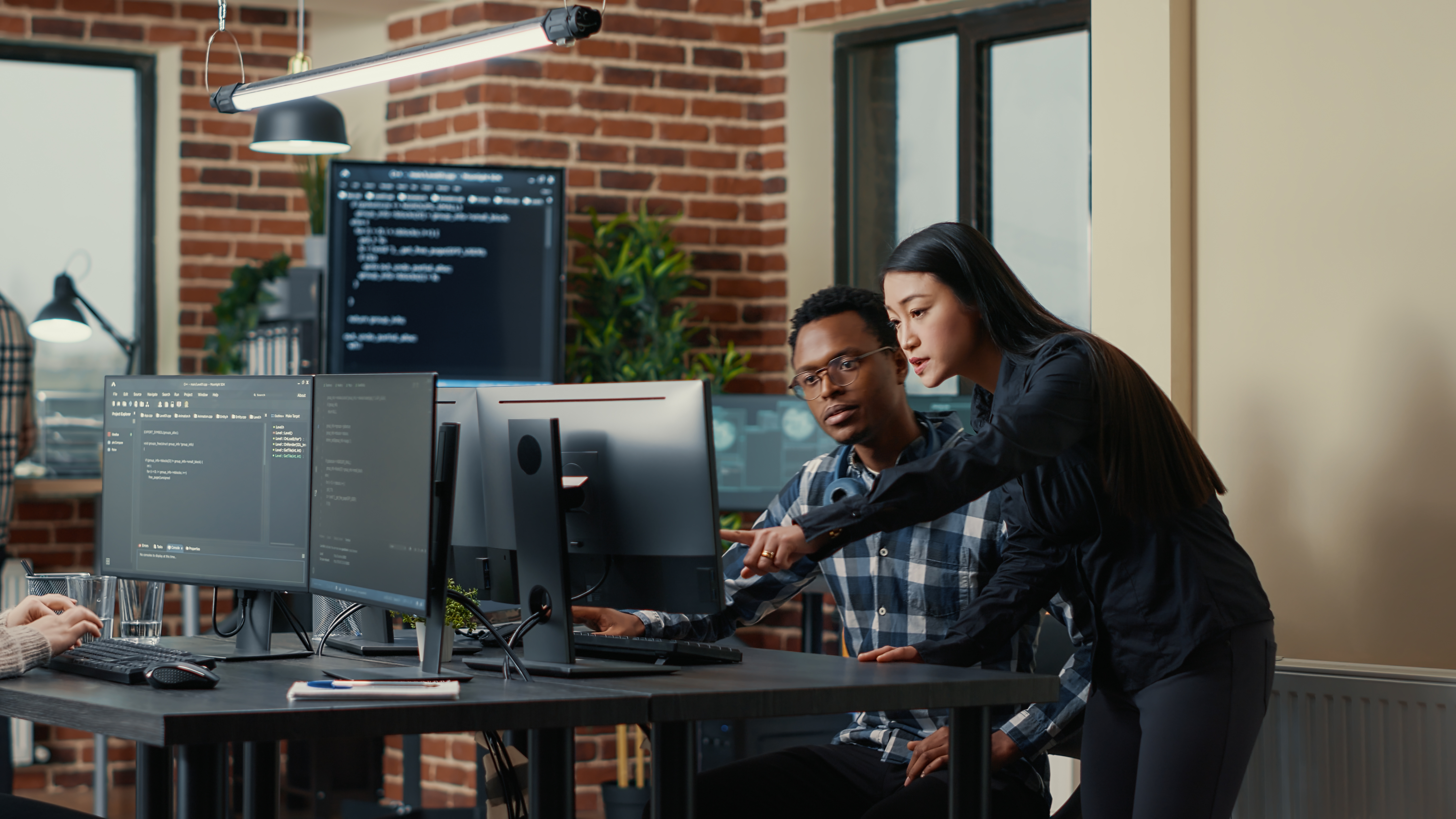 Two software developers holding laptop with coding interface walking towards desk and sitting down talking about group project. Programmers team discussing algorithms pointing at computer screen.